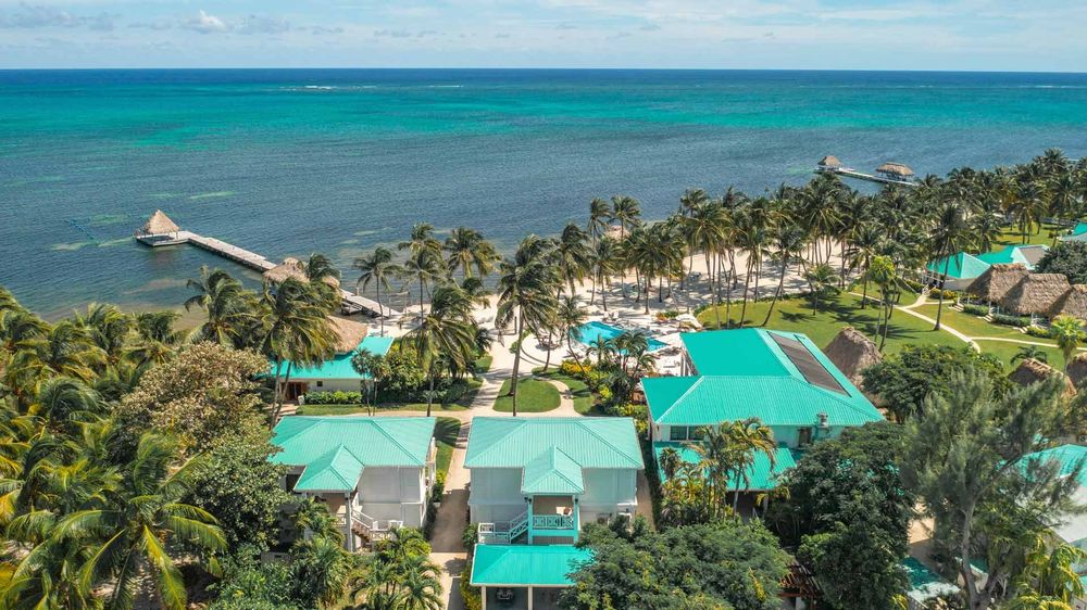 Aerial view of teal-roofed beach houses, palms and a pool along the shore of Ambergris Caye