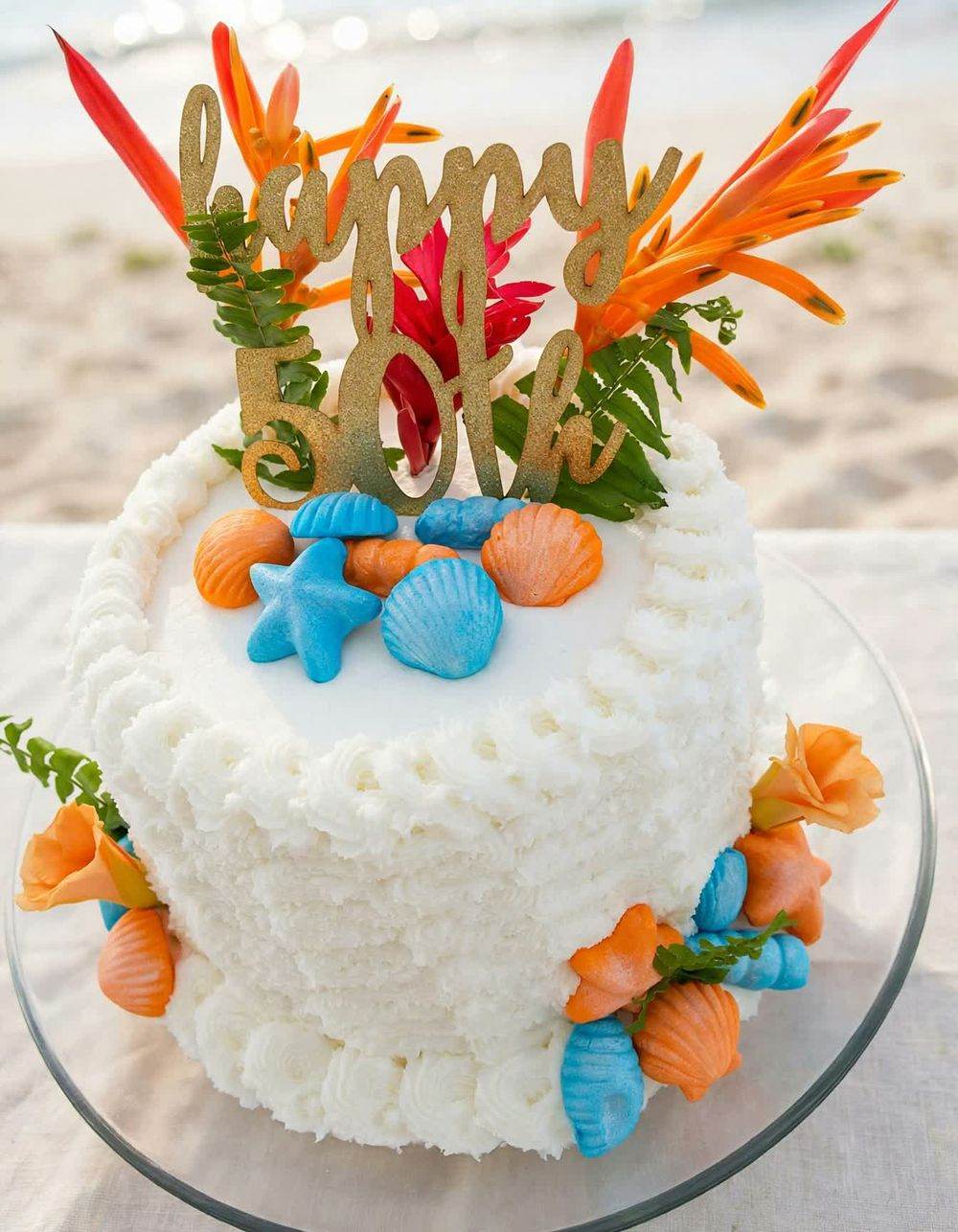 White 50th birthday cake decorated with seashells and tropical flowers, set on the beach